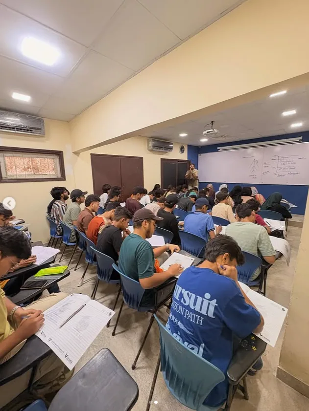 Students completing written exercises while a teacher explains a lesson at the whiteboard